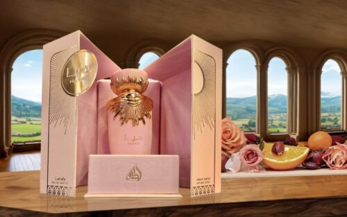 Botella de fragancia elegante en un estuche rosa, decorada con detalles dorados, rodeada de frutas y flores en un ambiente natural con ventanas arqueadas al fondo.