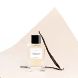 Frasco de Divine Vanille apoyado en un fondo de papel curvado color crema, con dos vainas de vainilla junto al frasco