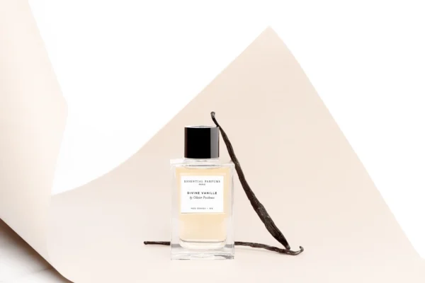 Frasco de Divine Vanille apoyado en un fondo de papel curvado color crema, con dos vainas de vainilla junto al frasco