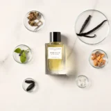 Frasco de Divine Vanille sobre mármol blanco entre placas de cristal con vainas de vainilla, haba tonka, resina de incienso, flores blancas y hojas