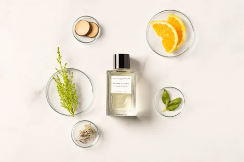 Essential Parfums Orange X Santal 100ml - Ingredients Frasco de Orange X Santal con naranja, hojas verdes, madera y musgo en composición cenital.