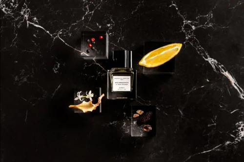 Essential Parfums Nice Bergamote Extrait 30 ml - Ingredientes Frasco de Nice Bergamote Extrait sobre mármol negro rodeado de medio limón, pimienta rosa, flores de azahar y haba tonka con resinas