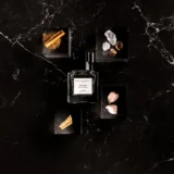 Frasco de The Musc Extrait sobre mármol negro rodeado de canela en rama, cristales de ámbar, pétalos de rosa y madera de sándalo