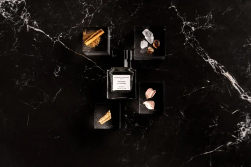 Frasco de The Musc Extrait sobre mármol negro rodeado de canela en rama, cristales de ámbar, pétalos de rosa y madera de sándalo